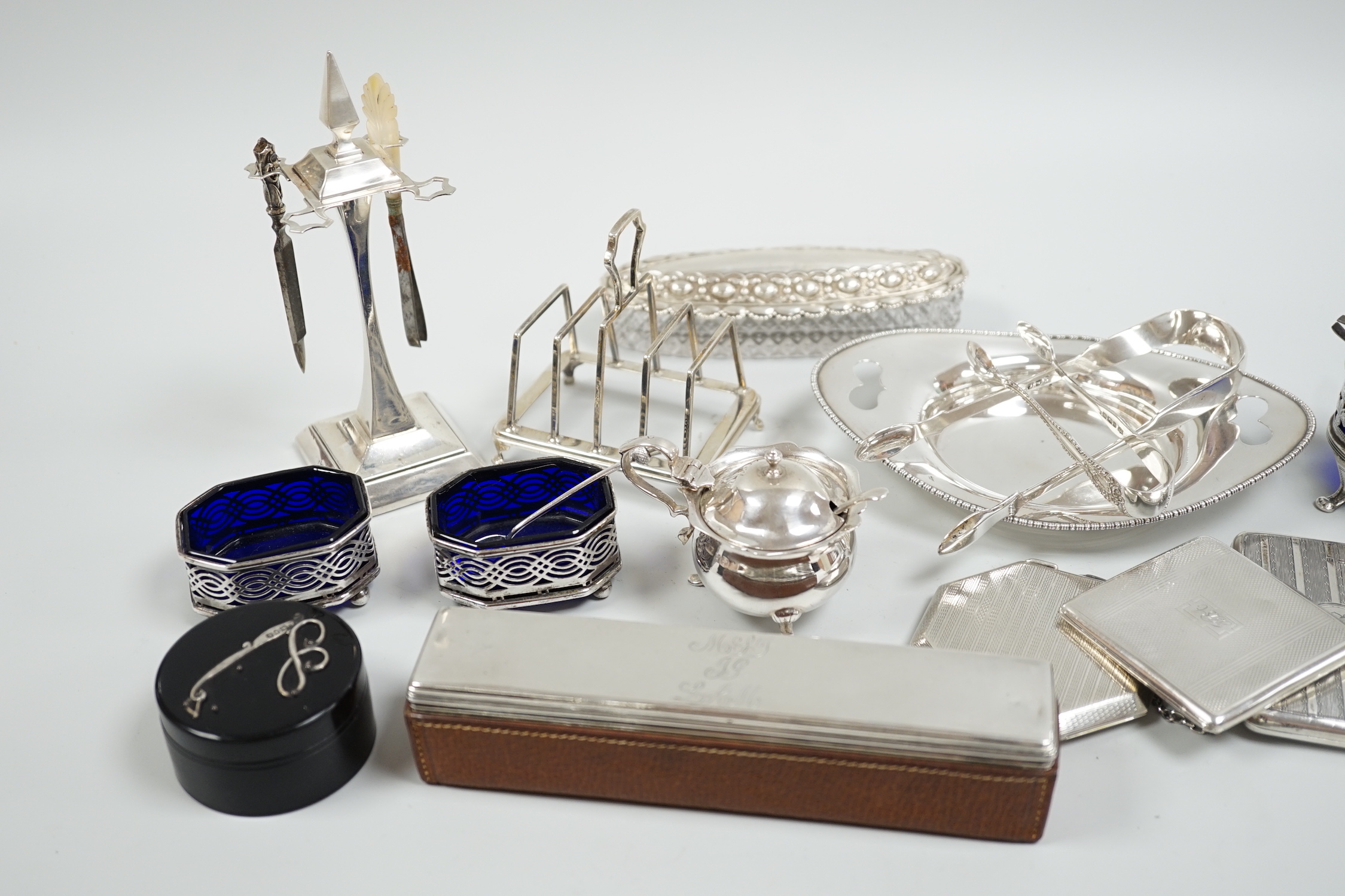 A group of mixed silver ware including a manicure stand, toastrack, purse, two compacts, four condiments, two pairs of sugar tongs, a stand and two mounted glass toilet jars.